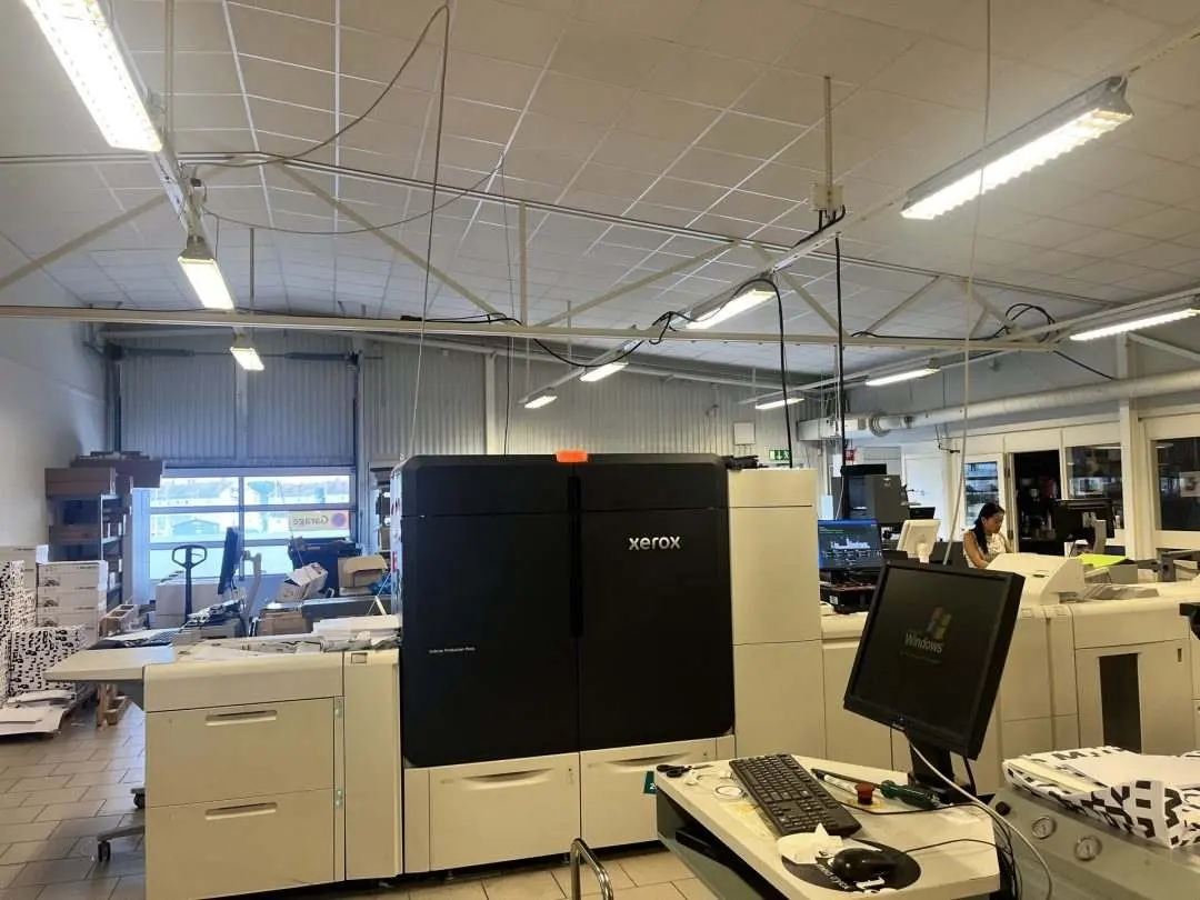A printing facility with various printing machines and equipment. The space is well-lit and organized, featuring a large black Xerox printer and a computer work
