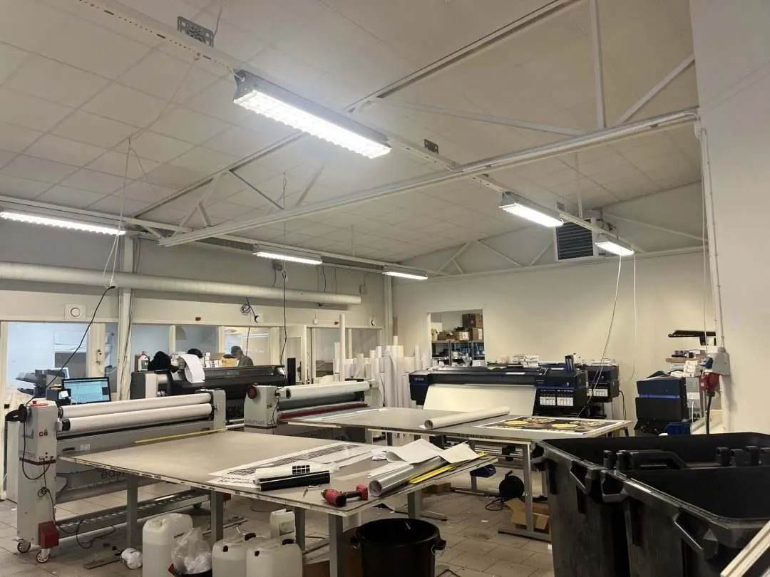 A spacious printing workshop with various machines and equipment. The room is well-lit with fluorescent lights and features large tables, printing devices, and 